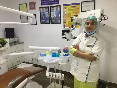A dentist in a white clinical coat and surgical cap standing with arms crossed beside a dental treatment chair and a ceiling-mounted dental microscope, certificates on the wall behind her.