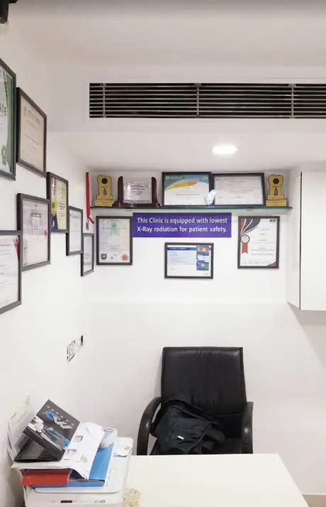 A corner of a consultation room with a shelf of trophies and certificates above a desk, a printer and a black office chair, and a wall sign about low-radiation X-ray safety.